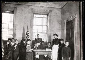 Around 10 schoolchildren of various races and genders line up with ballots in hand. One of two women teachers is placing a ballot in a box on a desk.