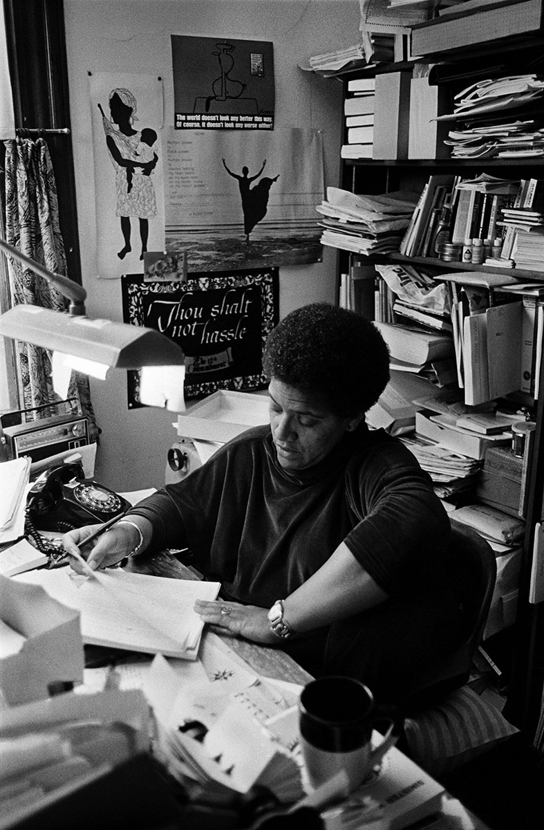 Audre Lorde, a Black woman, is seated at a desk in an office surrounded by books and papers. Her face tilts down toward a document she is reading.