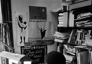 Audre Lorde, a Black woman, is seated at a desk in an office surrounded by books and papers. Her face tilts down toward a document she is reading.