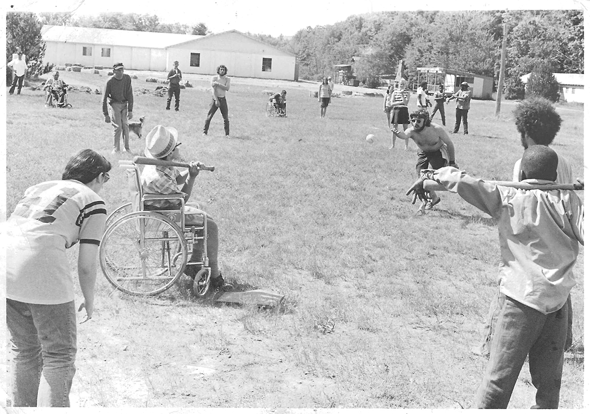 A young man pitches a ball to a boy sitting in a wheelchair and holding a bat. Other children and young adults look ready to react.