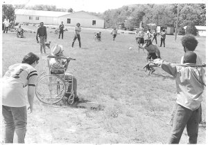 A young man pitches a ball to a boy sitting in a wheelchair and holding a bat. Other children and young adults look ready to react.