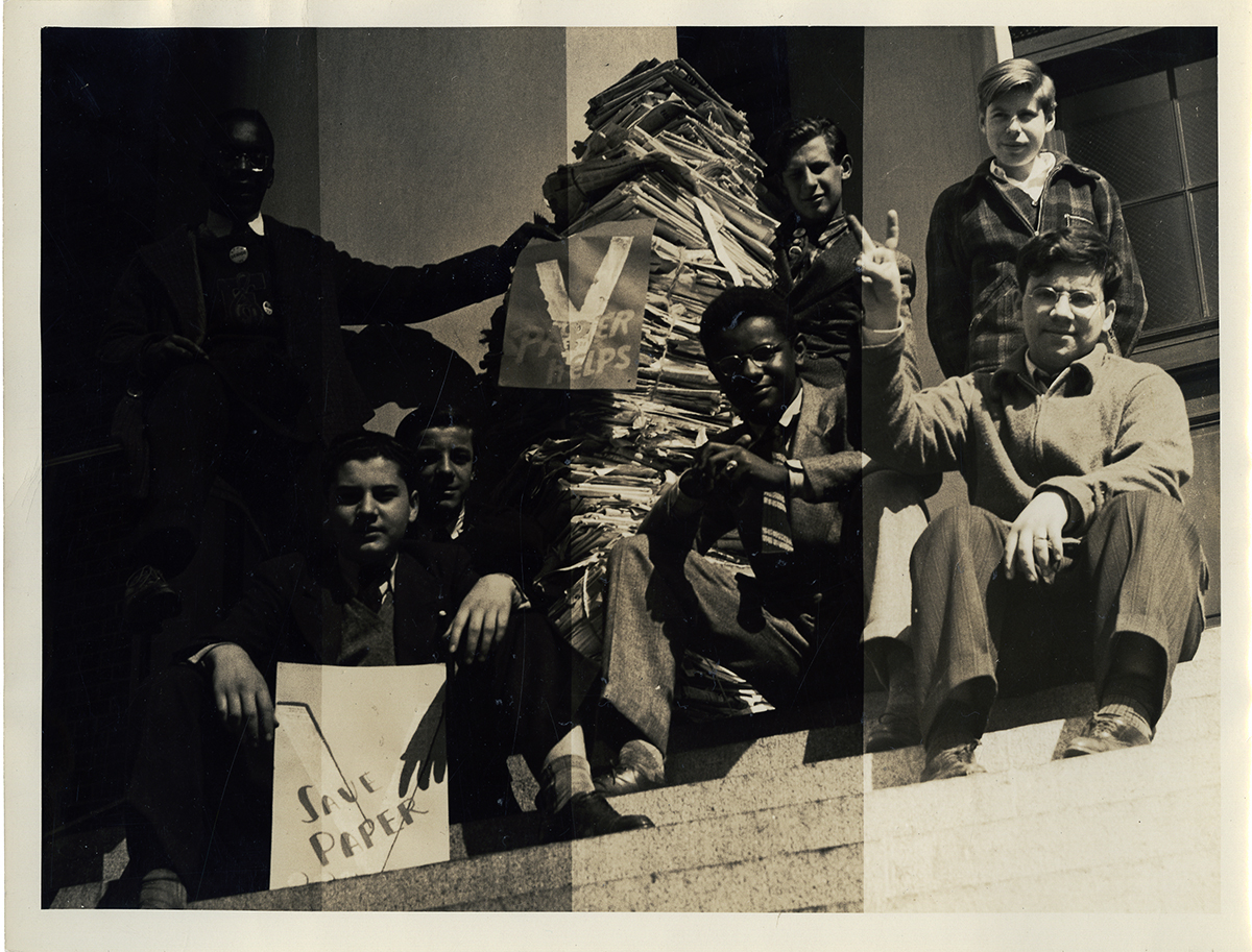 A group of Black and white children sit on steps beside a large stack of paper, making "V" signs with their hands and holding campaign signs