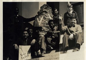 A group of Black and white children sit on steps beside a large stack of paper, making "V" signs with their hands and holding campaign signs