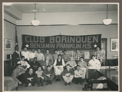 A group of Black and white children sit on steps beside a large stack of paper, making V signs with their hands and holding campaign signs.