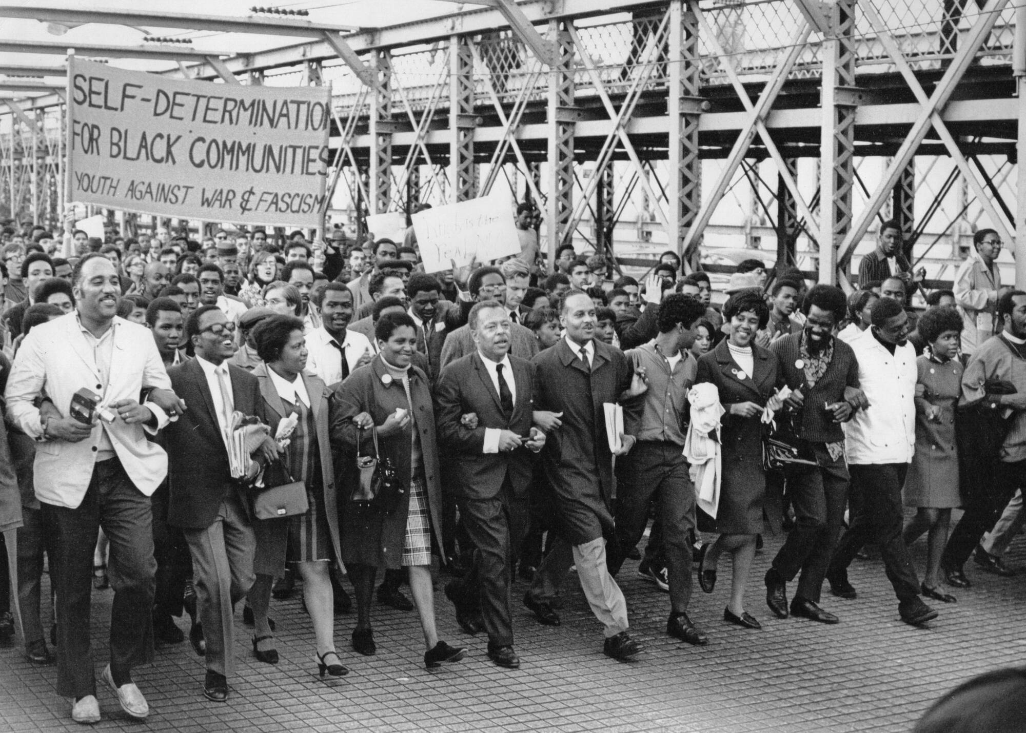 Black education advocates march over the Brooklyn Bridge together, arms linked. A large sign reads &quot;Self-Determination for Black Communities, Youth Against War & Fascism&quot;