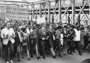 Black education advocates march over the Brooklyn Bridge together, arms linked. A large sign reads &quot;Self-Determination for Black Communities, Youth Against War & Fascism&quot;