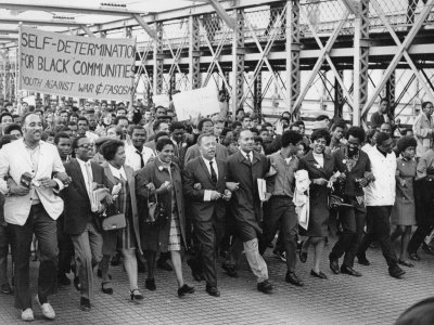 Black education advocates march over the Brooklyn Bridge together, arms linked. A large sign reads,"Self-Determination for Black Communities, Youth Against War & Fascism"