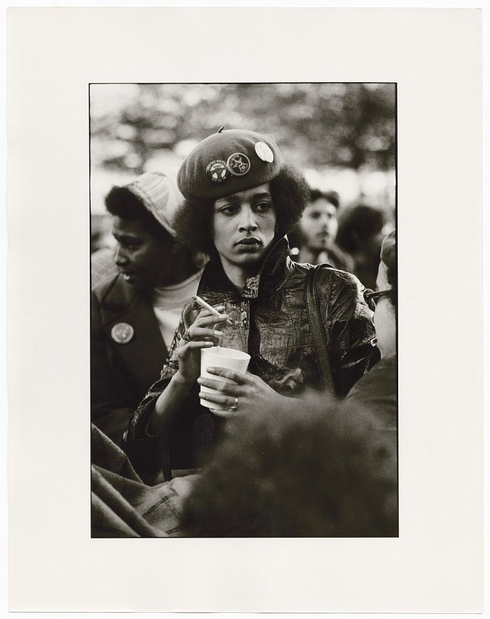 Denise Oliver, a young black woman, wearing a beret with several political buttons and a collared leather jacket. She's holding a cup and smoking a cigarette and has a somber expression on her face.