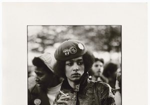 A young black woman with a somber expression wearing a beret with political buttons and a leather jacket. She's holding a disposable coffee cup and smoking a cigarette.