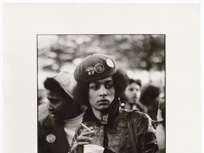 Denise Oliver, a young black woman, wearing a beret with several political buttons and a collared leather jacket. She's holding a cup and smoking a cigarette and has a somber expression on her face.