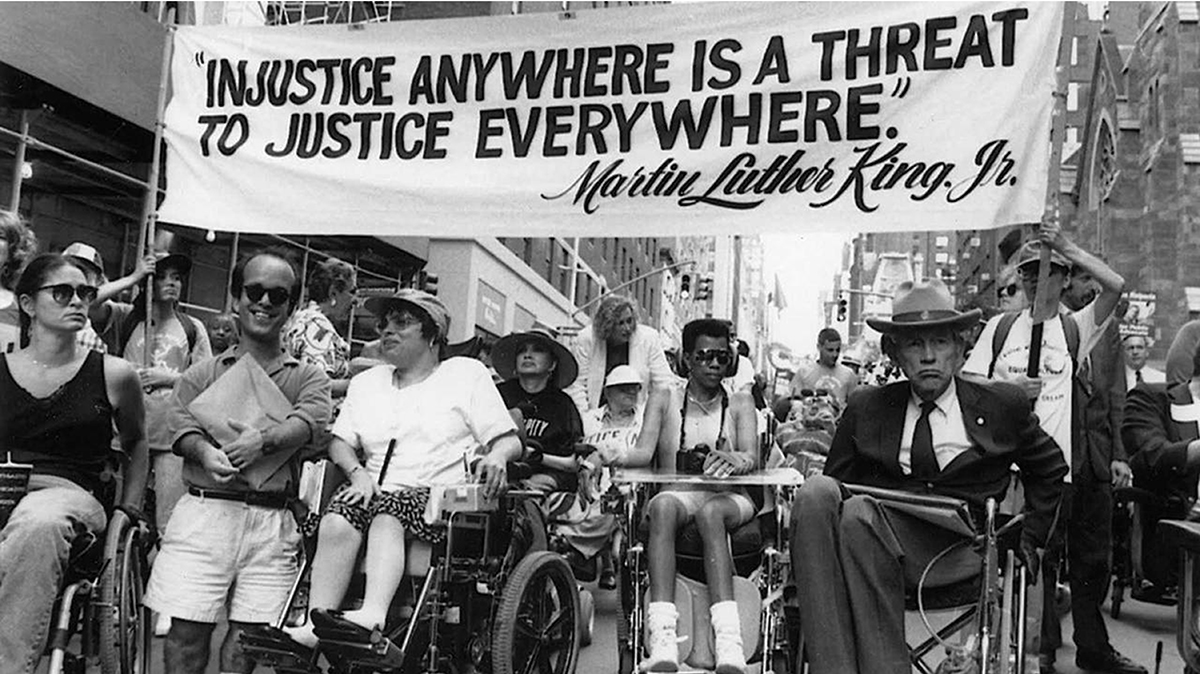 Several wheelchair users and a little person marchi in a parade with a banner