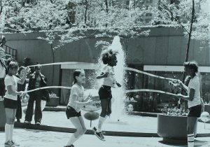 Girls jump rope near Lincoln Plaza. Two girls turn the ropes and the others jump. One girl is mid-air and a fountain behind her sprays upward. The other girl has knees bent, ready to jump.