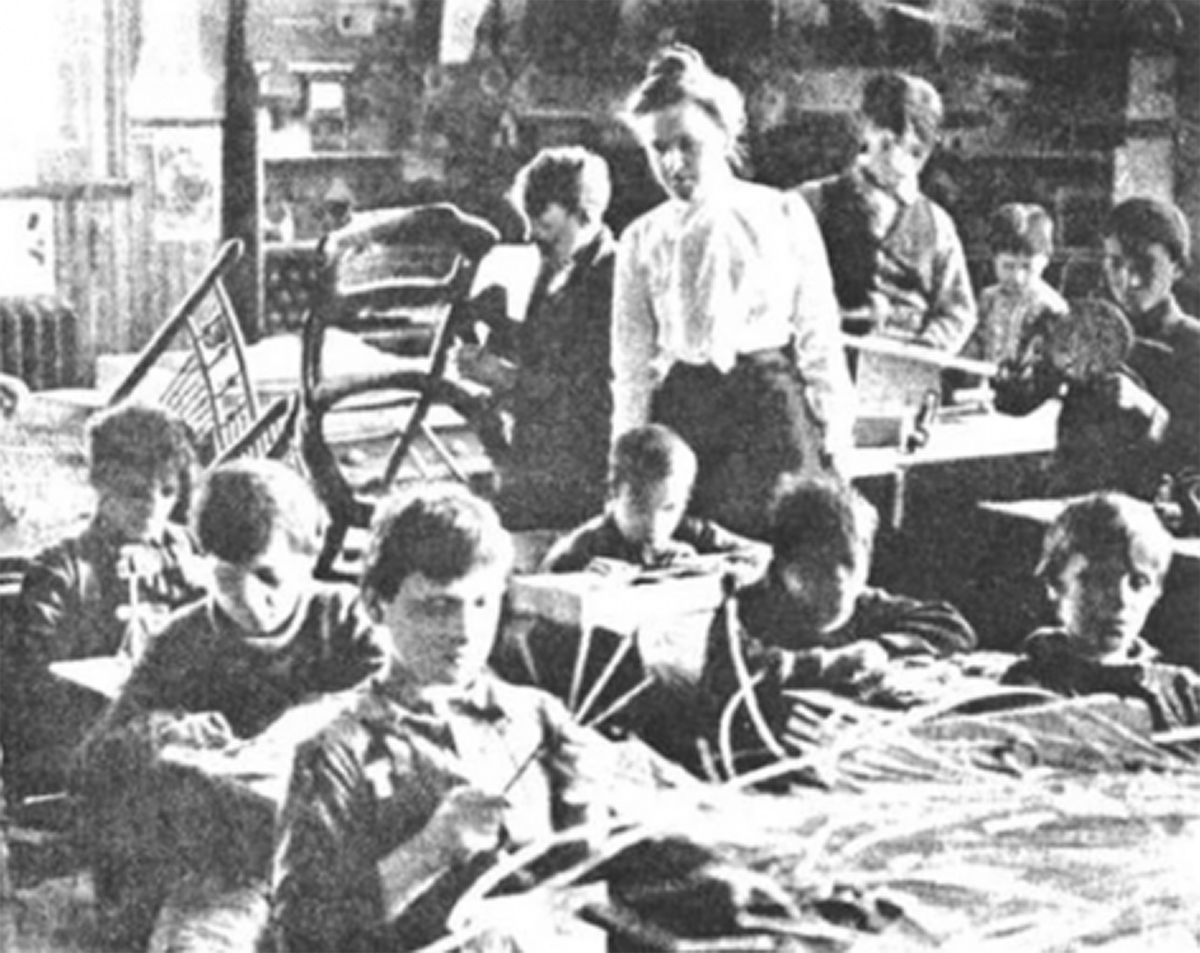 A white middle-aged woman teaches a classroom of boys of various ages making chairs