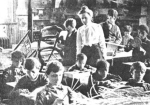 A white middle-aged woman teaches a classroom of boys of various ages making chairs