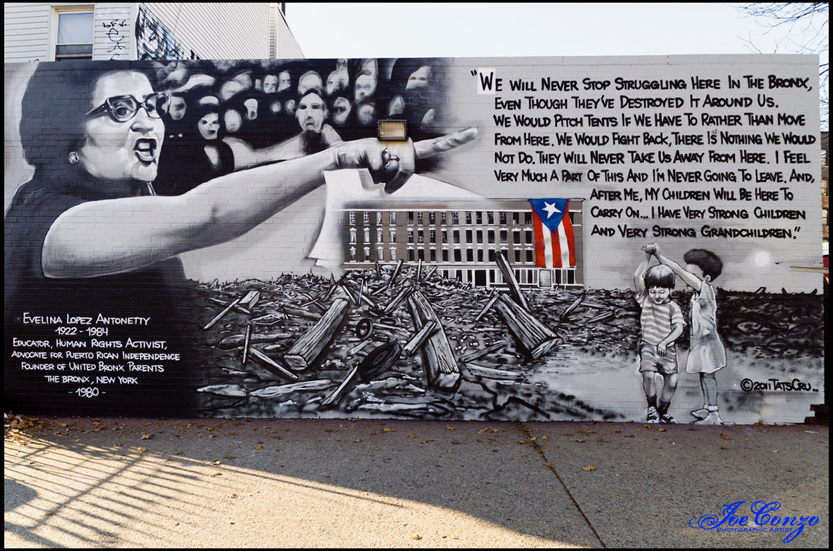 Black and white mural of Evelina Lopez Antonetty, a Puerto Rican woman, pointing with her left hand and speaking. The rest of the mural shows two children dancing in a field of rubble and a quote by Antonetty. A Puerto Rican flag hangs on a school building, the only colorful object in the painting.