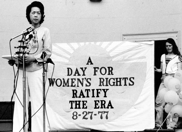 A Chinese American woman speaks into multiple microphones at a rally. A sign behind her reads &quot;A Day for Women's Rights. Ratify the ERA (Equal Rights Amendment) 8-27-77&quot;