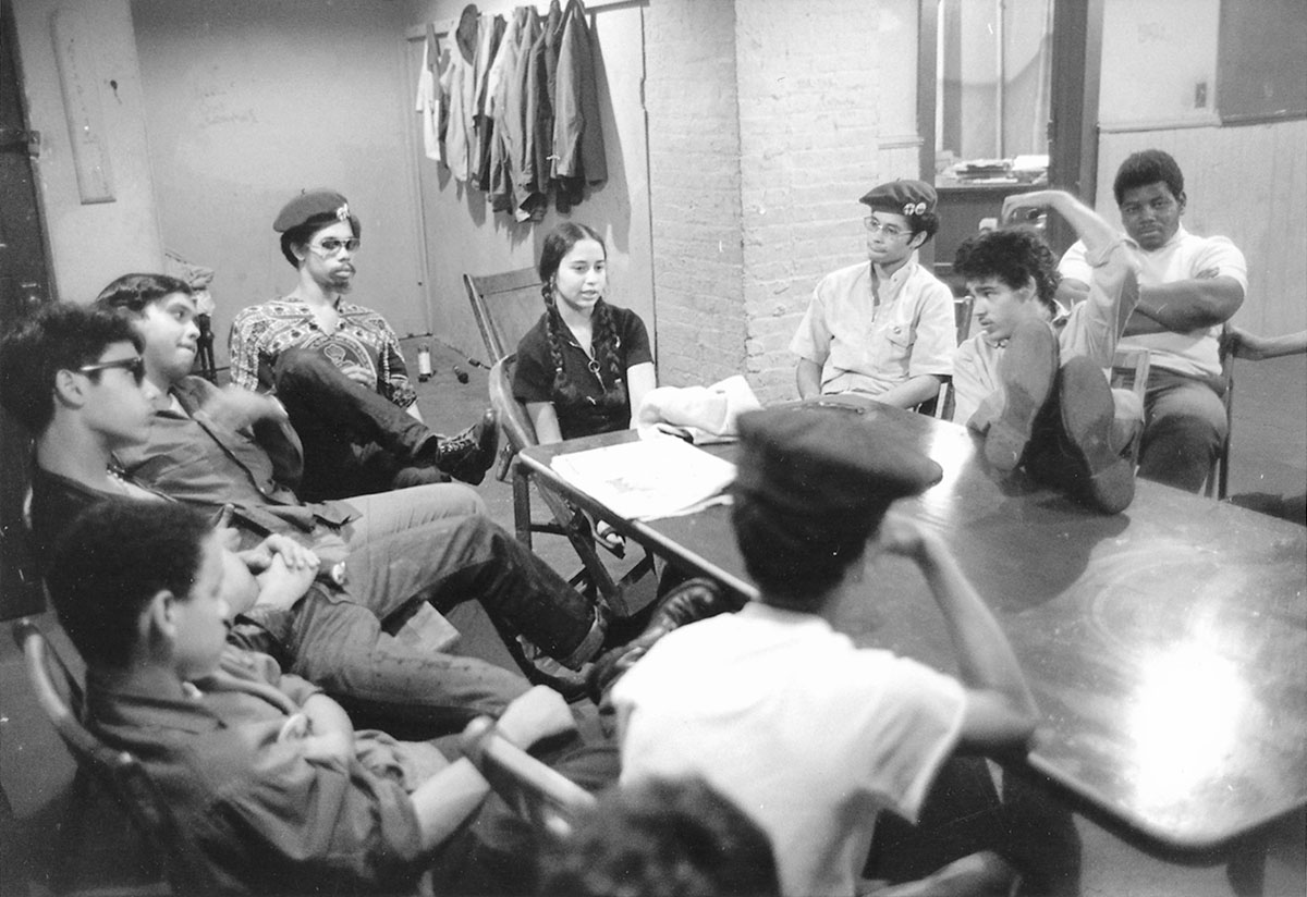 A group of young Black and Latino people are seated around a small table. Most appear to be male except for Iris Morales, who is at the head of the table.