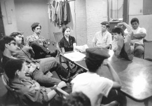 A group of young Black and Latino people are seated around a small table. Most appear to be male except for Iris Morales, who is at the head of the table.