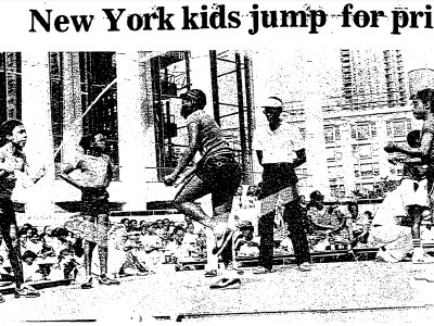 Black children jump rope in front of a crowd of seated people. A referree watches closely.