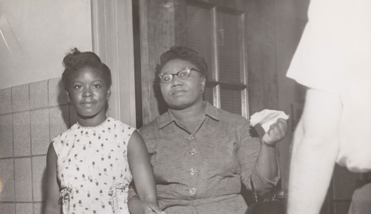 A middle-aged Black woman holds the arm of her teenaged daughter. They stand in a doorway, likely at a school.