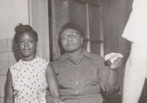 A middle-aged Black woman holds the arm of her teenaged daughter. They stand in a doorway, likely at a school.