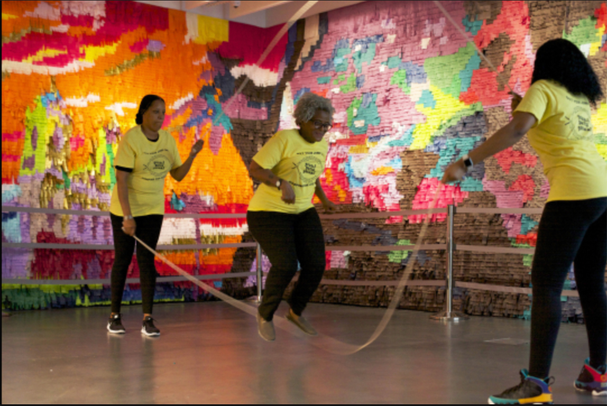 Reverand Malika Leigh Whitney, an older black woman, jumps rope with two women swinging two ropes.