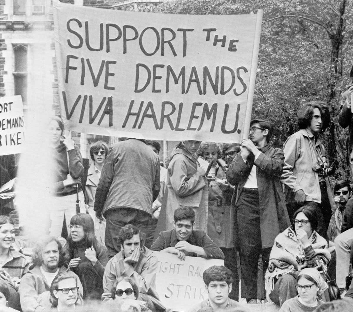 College students, most appearing to be white, hold signs at a rally outdoors with trees behind them. One student has a loudspeaker. The largest banner reads, "Support the Five Demands - Viva Harlem U."
