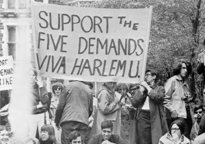 College students, most appearing to be white, hold signs at a rally outdoors with trees behind them. One student has a loudspeaker. The largest banner reads, "Support the Five Demands - Viva Harlem U."