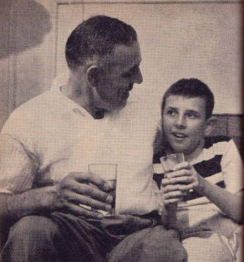 A white middle-aged man sits next to his son. They hold drinks and look at each other. The father has his arm around his son, and both are smiling.