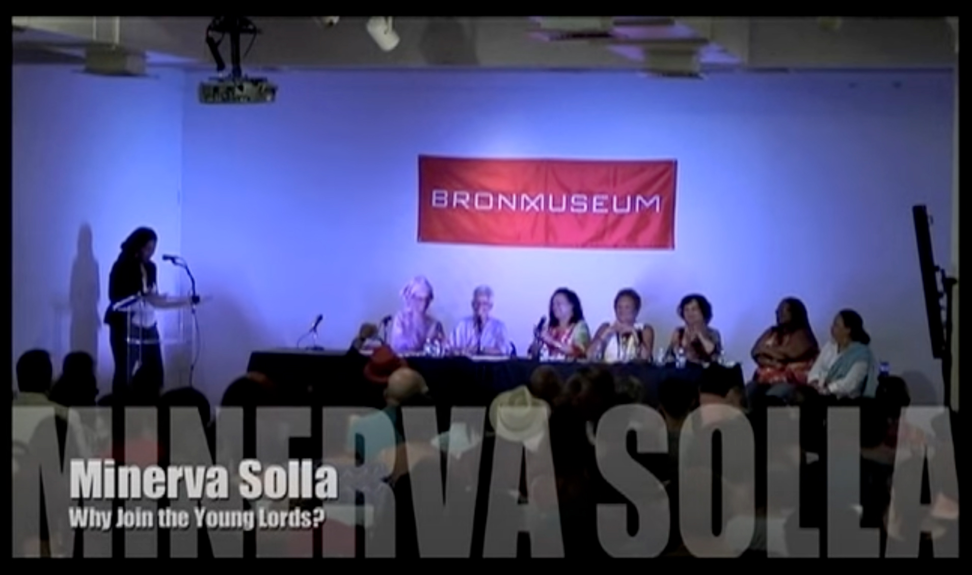 Still image from a videotaped event at the Bronx Museum showing a panel of women from the Young Lords sitting at a table and a speaker at a podium